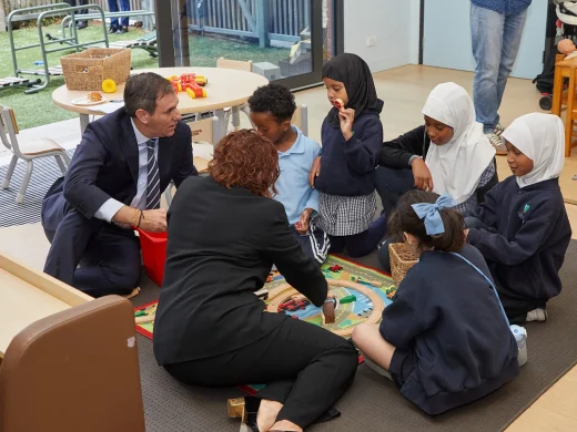 Two adults and five children sit on the floor around a wooden train set in a classroom, playing and talking together. The children wear school uniforms, and some wear white headscarves. A window shows a playground outside.