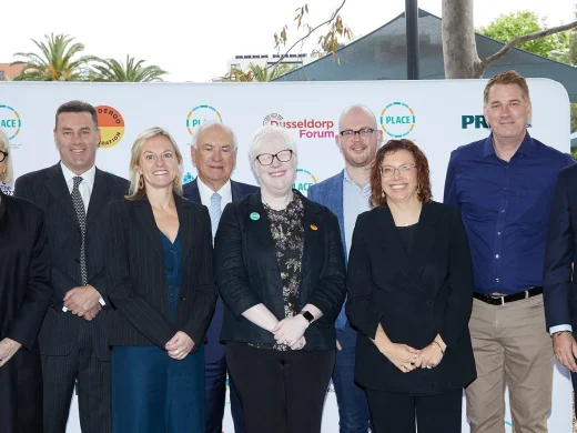 A group of nine adults, six men and three women, stand together outdoors, smiling at the camera in front of a branded backdrop with logos and greenery in the background.