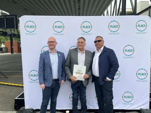Three men in jackets and jeans stand smiling in front of a banner with the I PLACE logo at an outdoor event. One man in the centre holds a folder. A tent roof and stage set-up are visible in the background.