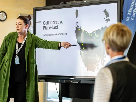 A woman in a green coat points at a digital map of Tasmania on a large screen displaying the title Collaborative Place-Led Catholic Schools in Tasmania, whilst another person looks on.