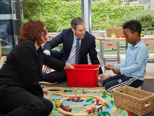 A man in a suit, a woman, and a young boy sit on the floor of a classroom, playing with wooden train tracks and toys. The man and woman smile at the boy, who is focused on a toy in his hands.