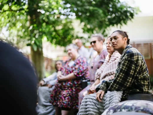 A group of people sit and listen attentively outdoors. One woman in a checked shirt is in focus, while others in colourful clothing sit on benches under leafy trees in the background.