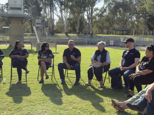 A group of people sit in a circle outdoors on a sunny day, engaging in conversation on a grassy area with trees and metal structures visible in the background.