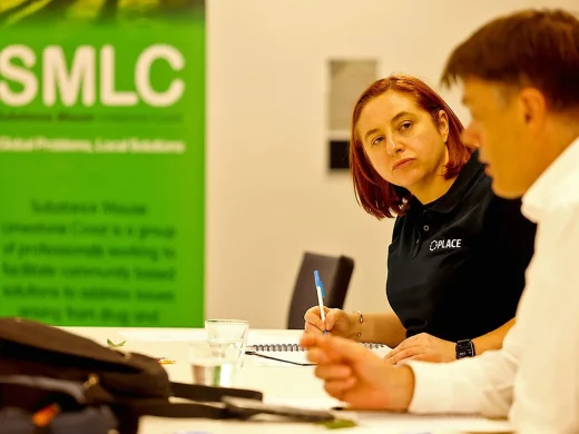 A woman with red hair listens attentively and takes notes during a discussion with a man at a table. A green SMLC banner is visible in the background.