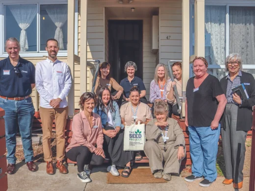 A group of thirteen smiling adults pose together on the steps and path outside a cream-coloured house, with some standing and others sitting. One person holds a tote bag labelled The Seed Project.