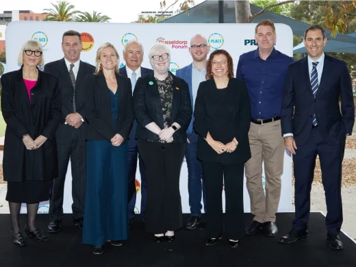 Nine adults in business attire stand together on a stage outdoors, smiling for a group photo in front of a white backdrop with colourful logos and text. Trees and buildings are visible in the background.