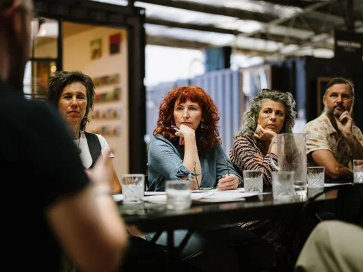 Four people sit at a table indoors, attentively listening to someone speaking off-camera. They appear engaged, with notebooks, papers, and glasses of water in front of them, suggesting a meeting or discussion.