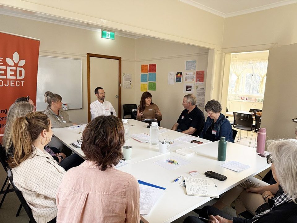A group of people sit around a large white table in a bright meeting room, engaged in discussion. Papers, notebooks, and drinks are on the table. A banner reading SEED PROJECT is visible on the left wall.