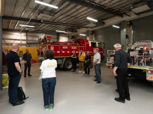 A group of people stands in a fire station garage, gathered around two parked fire engines. Some are listening to a speaker, and the space is bright with exposed ceilings and roller shutters.