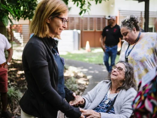 A woman stands holding hands with an older woman who is sitting in a wheelchair outdoors, surrounded by other people. The two women are making eye contact and sharing a meaningful moment.
