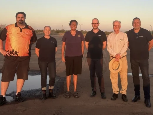 Six people stand side by side outdoors on a paved area at sunset, smiling at the camera. Some wear dark shirts with a logo, whilst one holds a straw hat. There are puddles and open space in the background.
