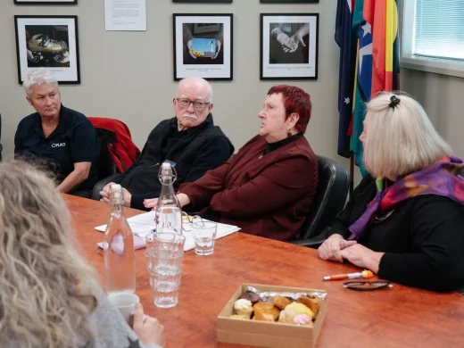 Five older adults sit around a conference table in discussion. There are pastries, cups, and water on the table. Framed photos and flags hang on the wall behind them. One woman appears to be speaking.