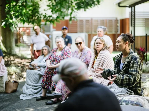 A group of people sit outdoors in a circle, some wrapped in blankets, listening to a woman speaking and gesturing with her hands. The setting is shaded by trees near a building.