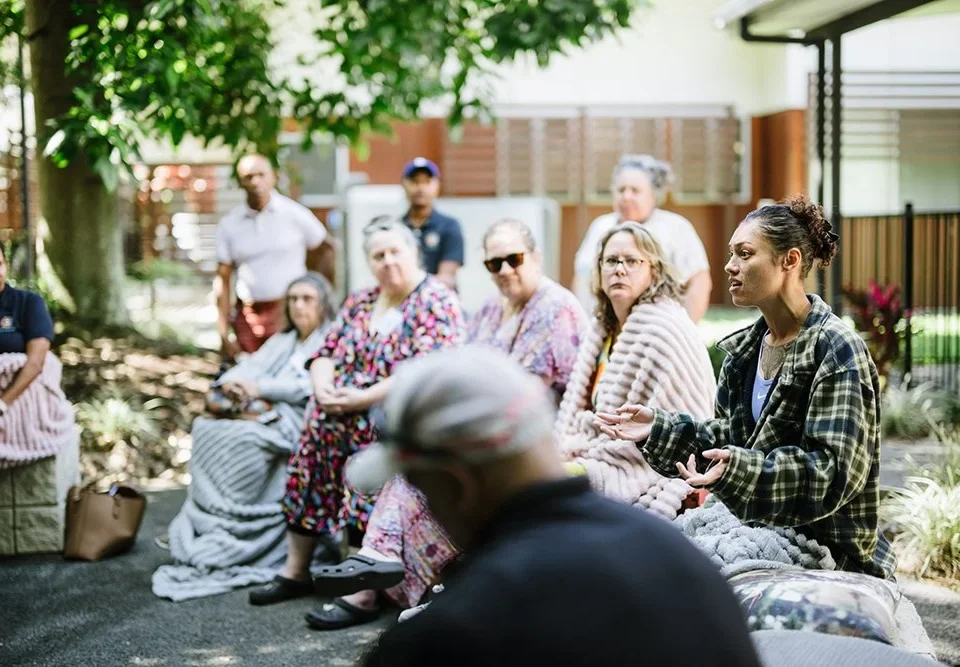 A group of people sit outdoors in a circle, some wrapped in blankets, listening to a woman speaking and gesturing with her hands. The setting is shaded by trees near a building.