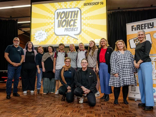 A group of people stand and kneel together, smiling for a photo in front of a large screen that reads Youth Voice Project Workshop 14. Posters and banners are visible on the stage behind them.