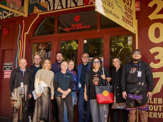 A group of eleven people stand smiling in front of Glebe Youth Service, outside a red and yellow building with the number 2037 painted on the wall. Some are holding papers, and one person wears an Aboriginal flag shirt.