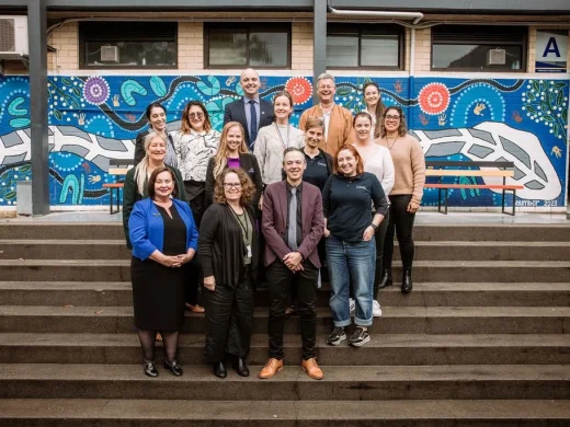 A group of sixteen adults stands and sits on outdoor steps in front of a colourful mural featuring abstract patterns and shapes. Most are smiling and appear to be posing for a group photo.