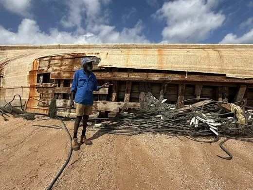 A person in a blue shirt, hat, and shorts stands on sandy ground beside a large, rusted shipwreck, with nets and ropes scattered nearby under a partly cloudy sky.