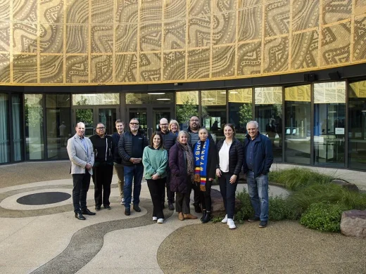 A group of twelve people stands outdoors in front of a modern building with glass doors and gold patterned panels; they are smiling, dressed in casual and semi-formal attire, and some are wearing scarves and jackets.