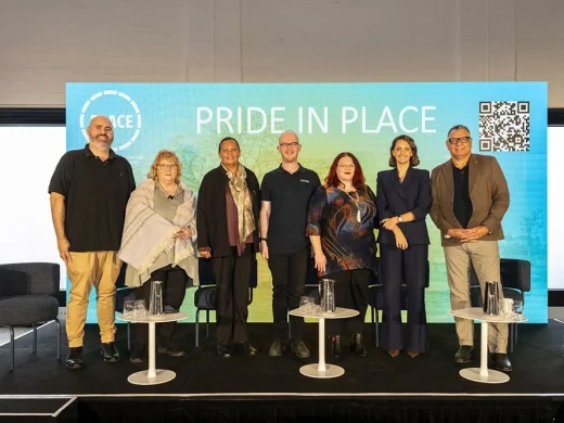 Seven people stand on a stage in front of a large screen displaying PRIDE IN PLACE with a QR code. Empty chairs and small tables with water glasses are in front of them. All are smiling and facing the camera.