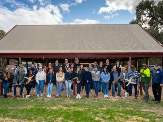 A group of about 30 people pose outdoors in front of a large building with a tin roof, smiling and holding boomerangs, with trees and a partly cloudy sky in the background.