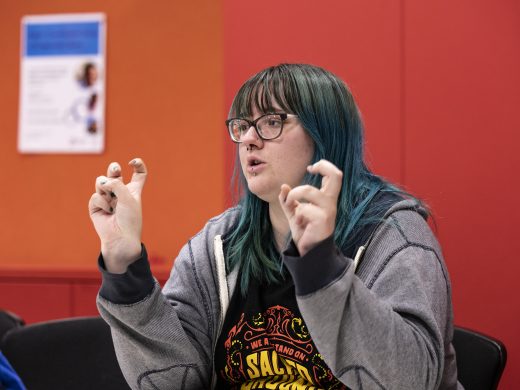 A person with long teal hair and glasses gestures with inverted commas while speaking, seated indoors against a red-orange background.