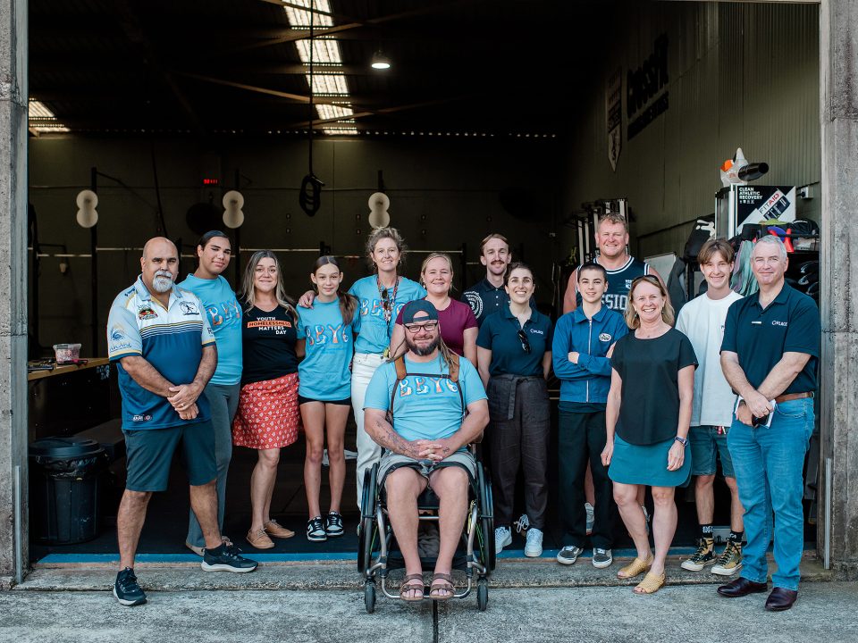 A diverse group of 15 adults and children smile and pose together inside a gymnasium, with one person in a wheelchair at the front centre. The gymnasium has equipment and weights visible in the background.