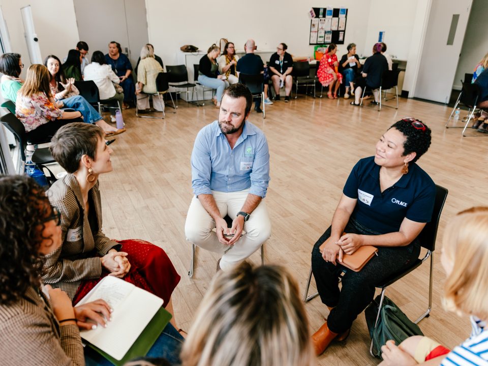 A group of adults sit in a circle having a discussion in a bright, spacious room, with other small groups talking in the background. Some people are taking notes and everyone appears engaged.