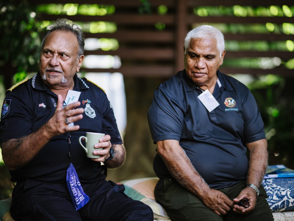 Two older men sit outdoors on cushions, engaged in conversation. One gestures while holding a mug; both wear dark shirts with embroidered logos and name badges. Greenery and wooden fencing are visible in the background.