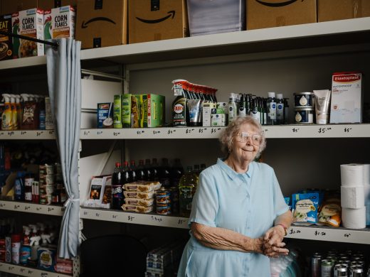 An elderly woman in a light blue dress stands in front of stocked shelves in a small shop, surrounded by food, cleaning products, and bottled drinks. Large cardboard boxes with Amazon logos are stacked above the shelves.