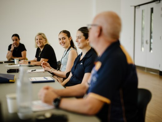 Five people sit at a long table in a bright meeting room, engaged in conversation and smiling. Papers, cups, and a water bottle are on the table. The background shows white walls and double doors.