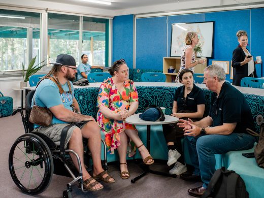 A group of four adults, including a man in a wheelchair, sit and talk around a small table in a brightly lit, modern room. Two women stand in the background, one holding a drink and smiling.