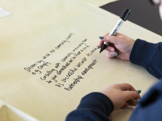 A person writes notes with a black marker on a large sheet of paper. The notes mention topics like community sport, consulting with communities, and partnership development. Only the person's hands and part of their arm are visible.