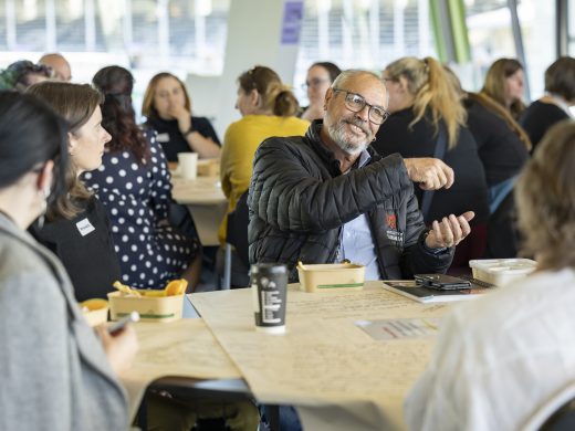 A group of people sit around tables in discussion at a workshop or meeting. A man in glasses gestures as he speaks whilst others listen, with food containers, drinks, and papers on the table.