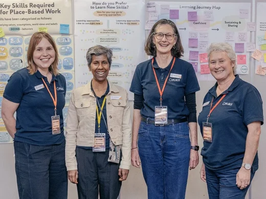 Four smiling women wearing navy OPAKE shirts and conference lanyards stand in front of colourful posters about learning skills and journey mapping, featuring diagrams and sticky notes.