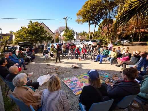 A diverse group of people sit in a large outdoor circle on chairs, attentively listening to a speaker in the centre. Colourful artworks are laid on the ground, and trees and houses surround the gathering on a sunny day.