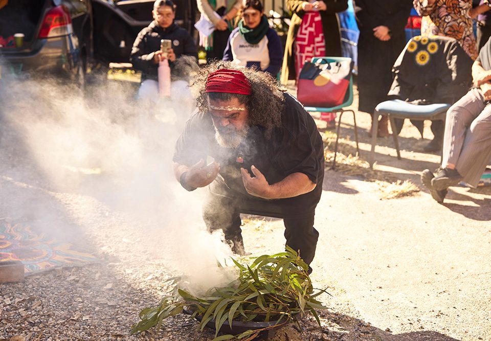 A man with long hair and a red headband bends over a smoking ceremonial fire, holding his hands up. People sit and stand in the background, watching the outdoor event under bright sunlight.