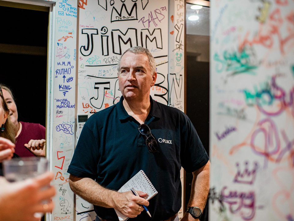 A man in a black polo shirt holding a notepad stands in front of a wall covered with colourful signatures and doodles. People are partially visible around him, and he appears to be listening intently.