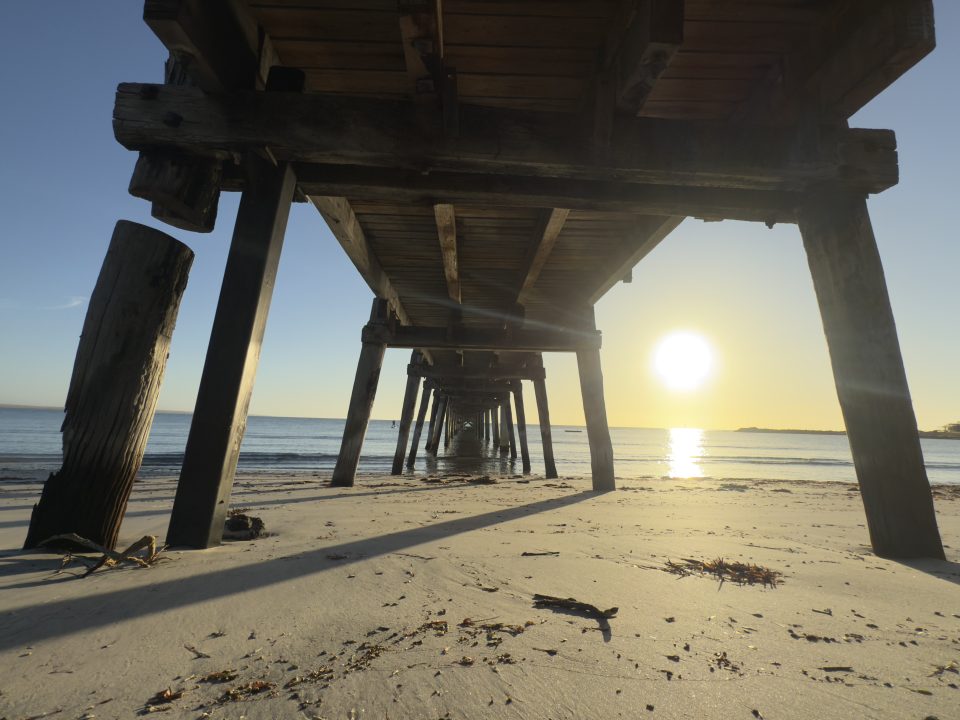 View from under a wooden pier on a sandy beach at sunset, with the sun low in the sky over the sea, casting long shadows and illuminating the sand and water.