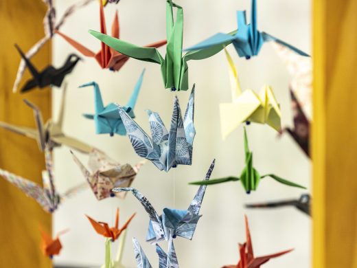 Colourful paper origami cranes hang from strings, creating a vibrant and delicate mobile against a blurred background. The cranes are made from various patterned and solid-coloured papers.