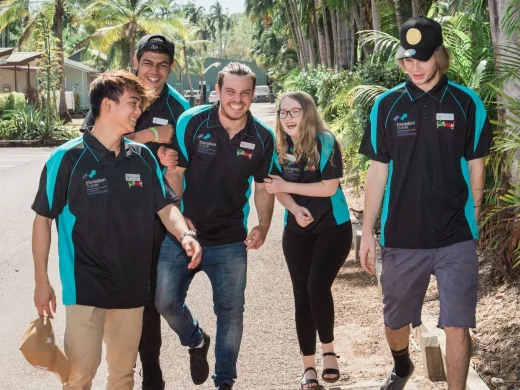 Five young adults, wearing matching black and teal shirts, walk and laugh together on a sunny path lined with palm trees. Two hold brown paper bags, and the group appears cheerful and relaxed.