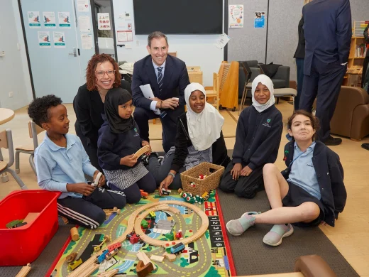 A group of children sit and play with wooden train tracks on a colourful mat, while two adults kneel behind them smiling in a classroom setting. The atmosphere is friendly and engaging.