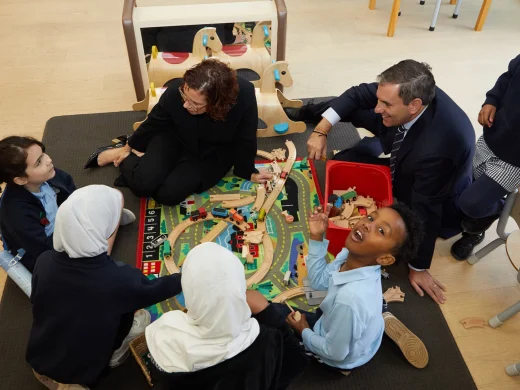 Six children and two adults sit round a table playing with a wooden train set and toy tracks. The group is engaged and smiling in a classroom or playroom setting.