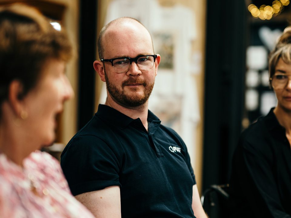 A man with glasses and a dark polo shirt sits indoors, looking attentively at a woman speaking in the foreground. Another woman is partially visible to his right. The setting appears casual and social.