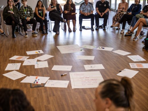 A group of people sit in a circle on chairs in a spacious room. In the centre of the circle, large sheets of paper with handwritten notes and markers are spread out on the wooden floor.