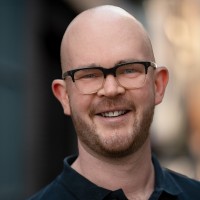 Luke is wearing a navy polo and black-framed glasses, smiling at the camera.