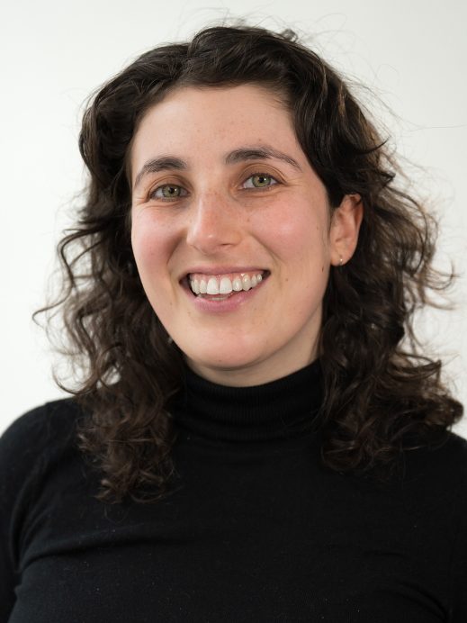 A woman with curly brown hair, wearing a black polo-neck, smiles at the camera against a plain white background.
