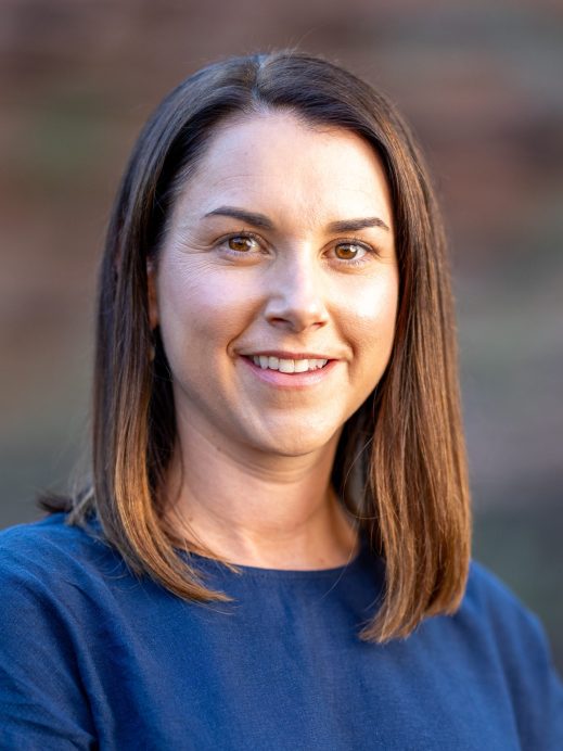 A woman with straight brown hair, wearing a navy blue top, smiles at the camera in an outdoor setting with a blurred brown and green background.