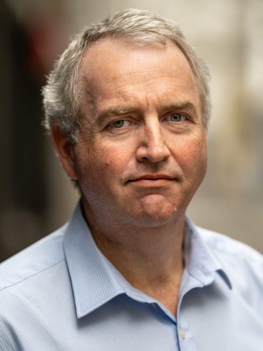 A middle-aged man with short grey hair and blue eyes, wearing a light blue collared shirt, stands outside with a neutral expression. The background is blurred.