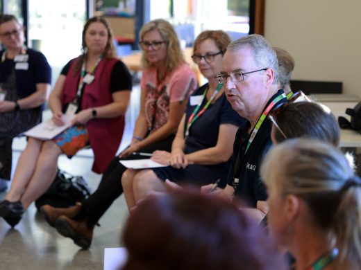 A group of adults sit in a circle indoors, attentively listening to a man speaking. They hold papers, suggesting a discussion or workshop setting. The atmosphere appears focused and engaged.